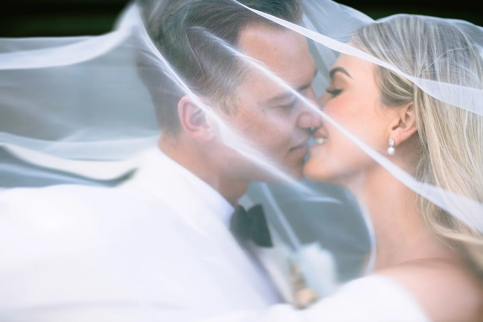 Extreme close-up of the bride and groom kissing beneath a sweeping tulle veil, her pearl drop earring and his black bow tie visible through the sheer fabric.