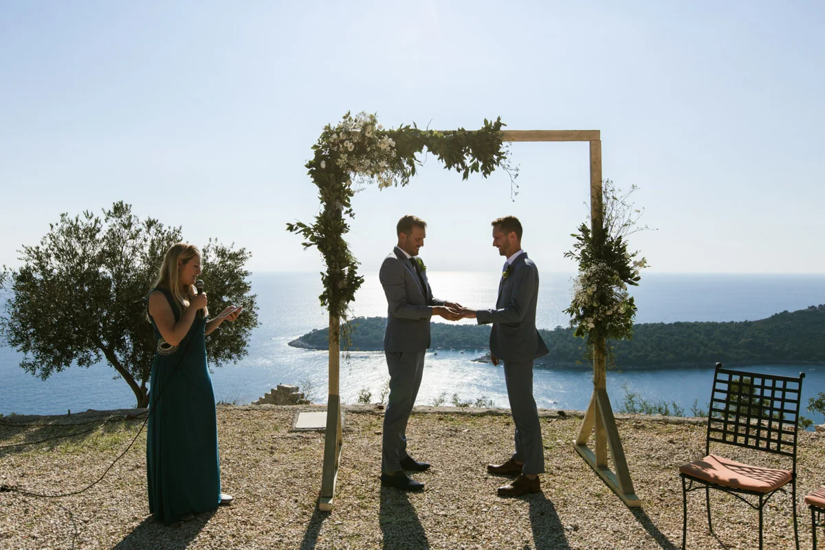 Two grooms exchange vows beneath the flower arch at Park Orsula above the sea