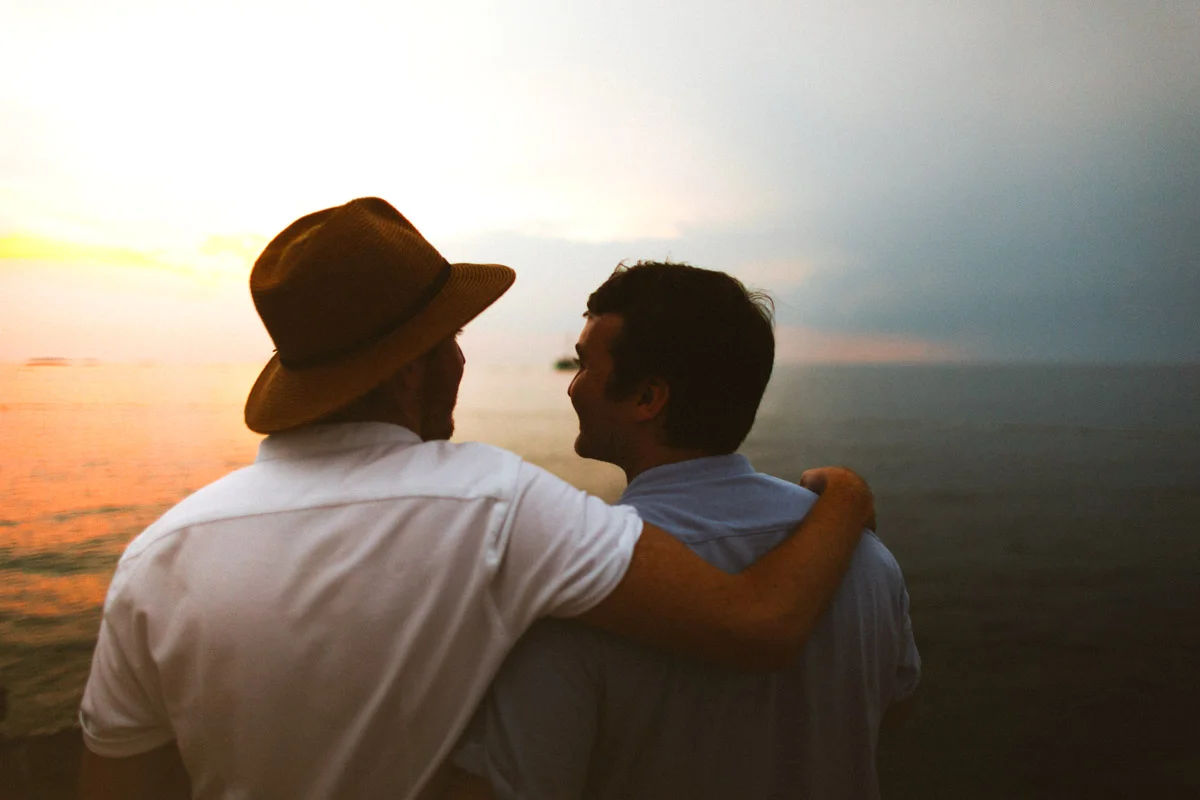 Two grooms embrace at water edge under orange sunset sky over calm Adriatic