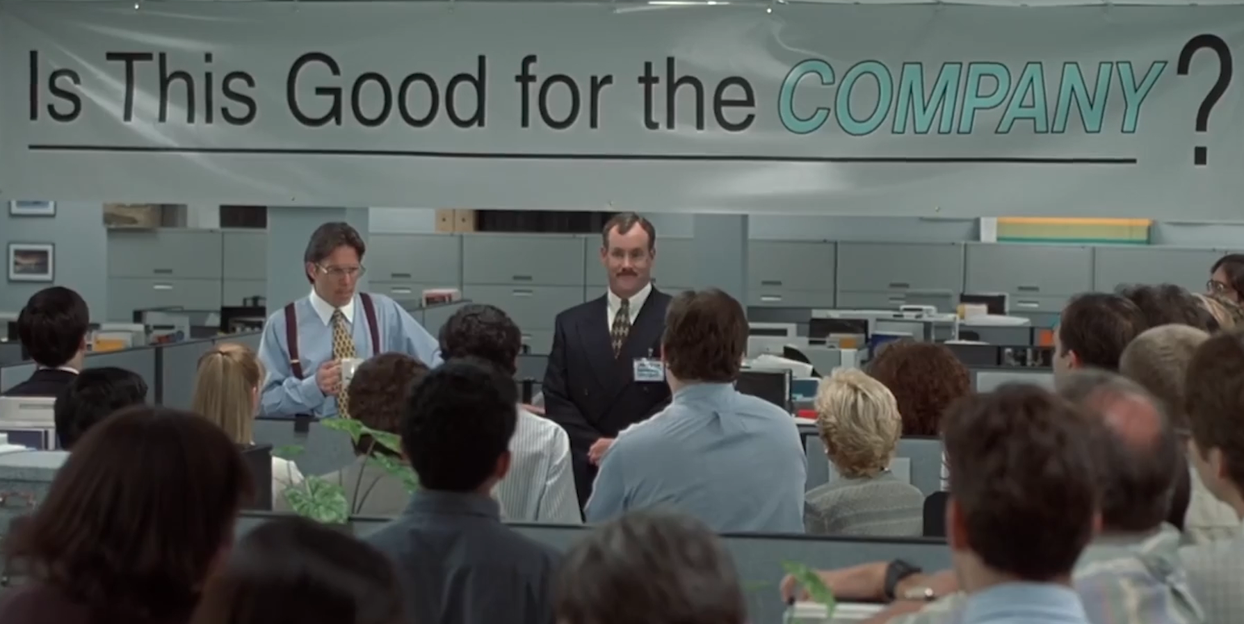 Office Space movie scene with a corporate meeting banner asking 'Is This Good for the Company?' highlighting corporate focus on efficiency.