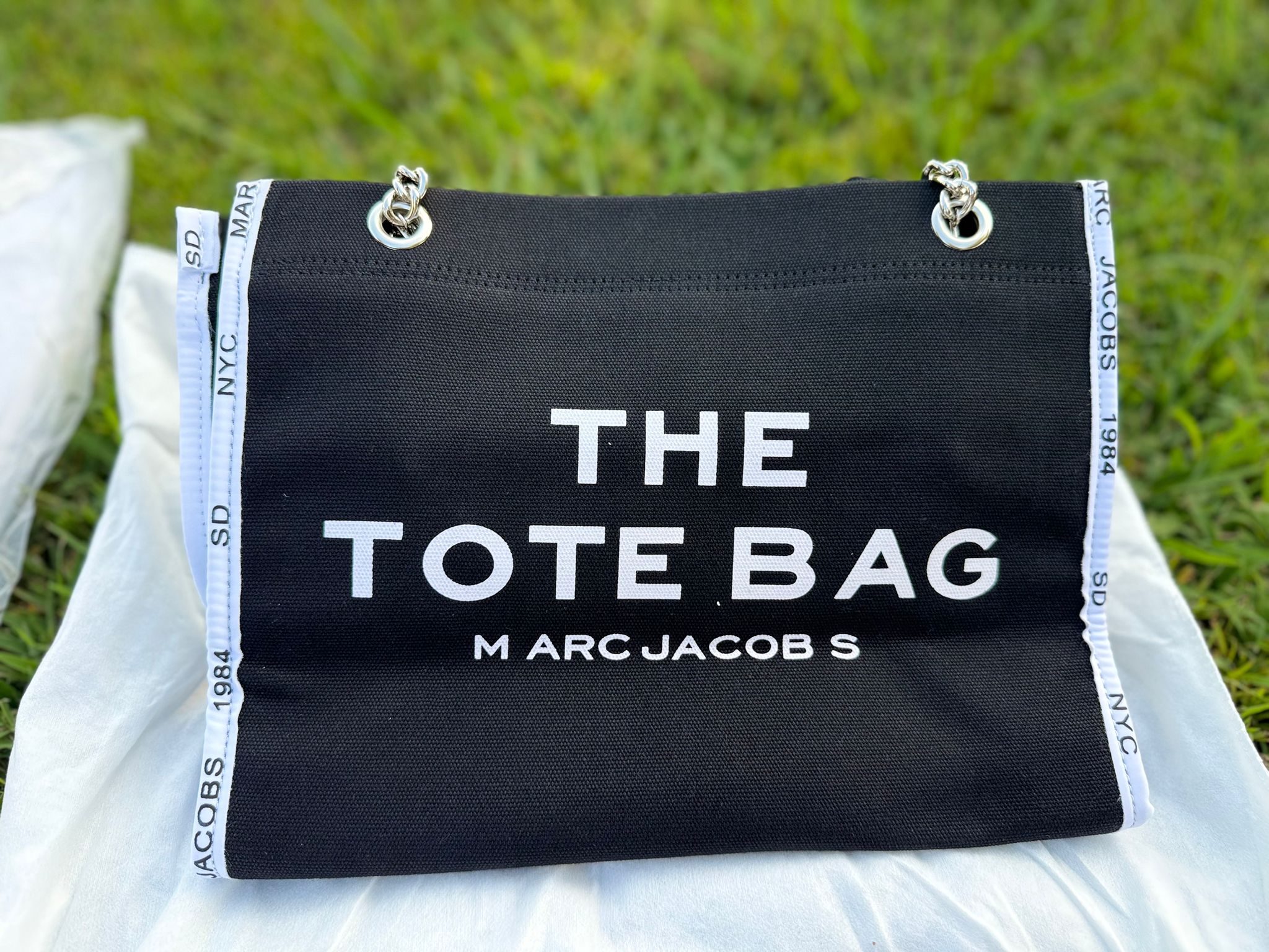 Make bold moves with the Marc-Inspired Statement Tote – a high-fashion essential that blends minimalism with edge. Crafted from durable black canvas, this spacious tote features sharp white side tape with signature city prints and a chunky silver-tone chain strap for that luxe finish. Ideal for everyday errands, airport style, or your favorite casual fits – this bag turns every outfit into a look. Lightweight, roomy, and effortlessly cool.