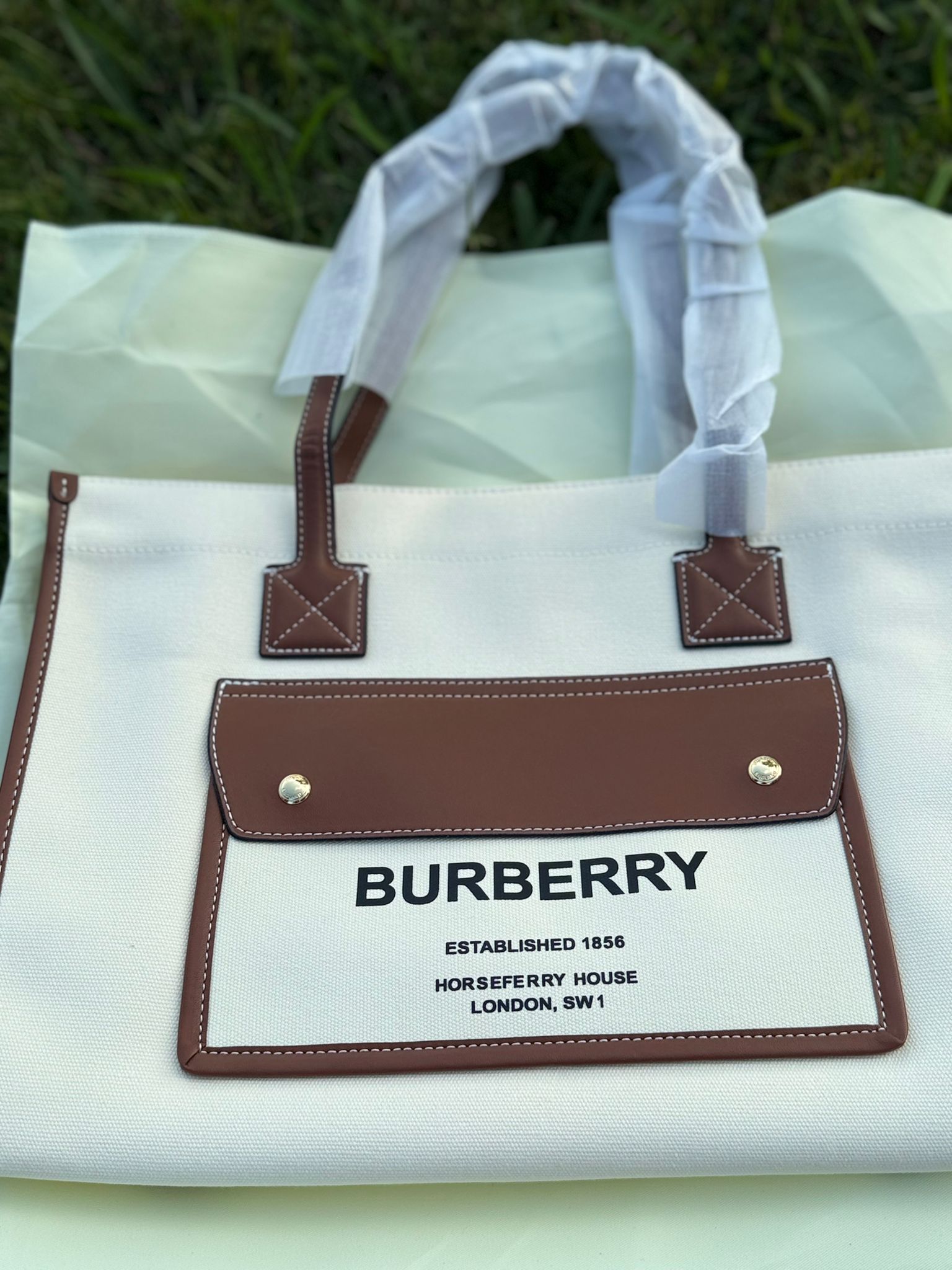 Embrace iconic British design with this sophisticated Burberry Horseferry Tote Bag. Crafted in sturdy off-white canvas and trimmed with luxurious tan leather, this bag features bold black text detailing and practical structure. It's perfect for everyday use—combining utility, comfort, and timeless style in one piece. Durable cotton canvas body with contrast leather trims Exterior flap pocket with BURBERRY logo and Horseferry House print Double shoulder handles with reinforced stitching Spacious open-top design Lightweight yet roomy for everyday carry 