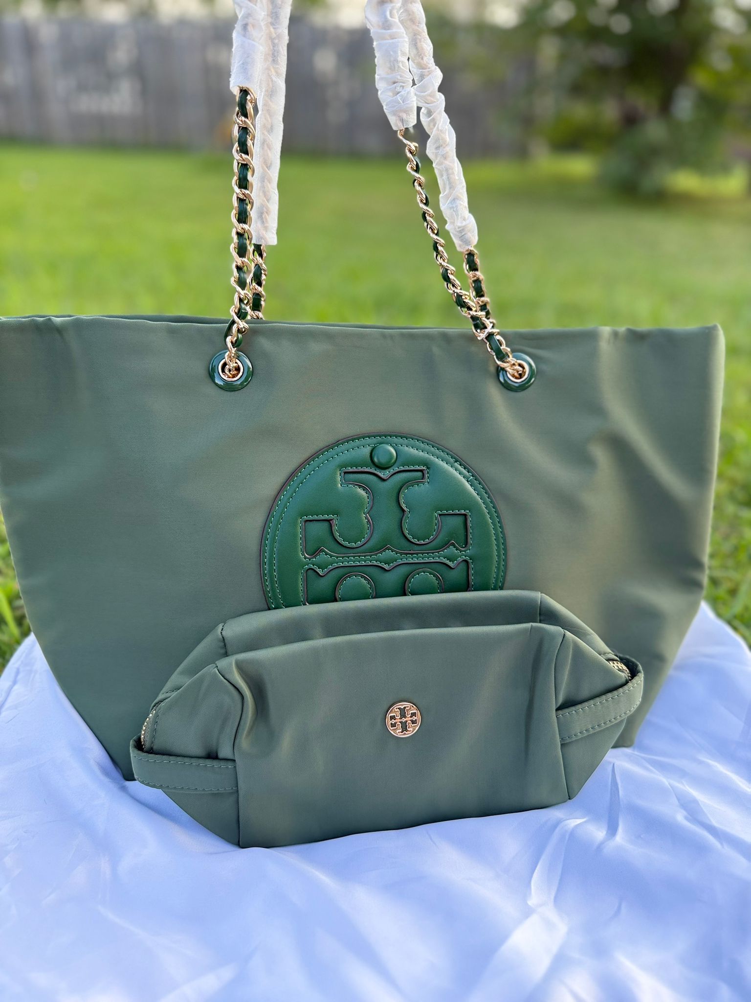 Sleek, functional, and unmistakably chic — this Tory Burch nylon tote bag comes in a lush forest green shade with a bold embossed logo at the front. It includes a matching detachable pouch that snaps shut with a signature gold-tone logo button. Perfect for everyday errands, work, or travel, this bag delivers effortless style and organization. Durable water-resistant nylon body Signature oversized Tory Burch logo in matching green leather Gold-tone hardware and chain-wrapped shoulder straps Includes matching zip pouch with gold button snap closure Spacious interior to fit daily essentials and more