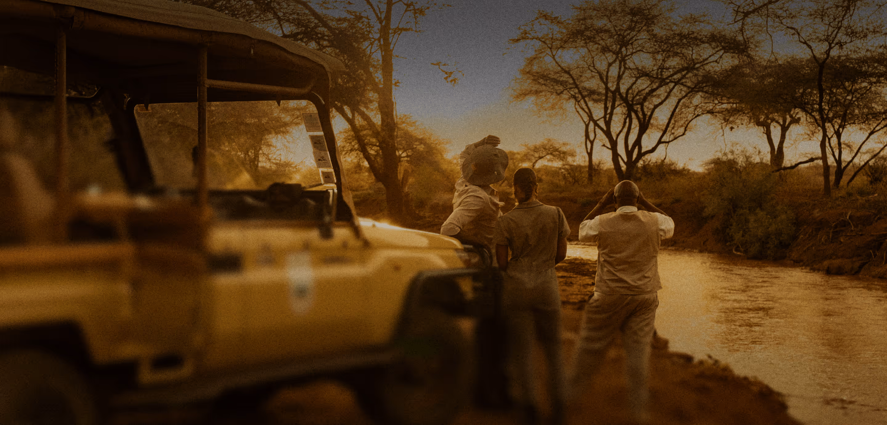 Drei Personen bei einer Safari beobachten einen Fluss am Abend mit einem Jeep neben ihnen.