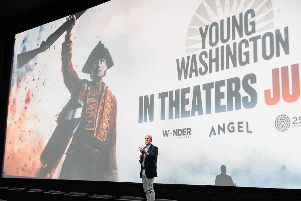 Man speaking on stage in front of a large movie poster for Young Washington in theaters.