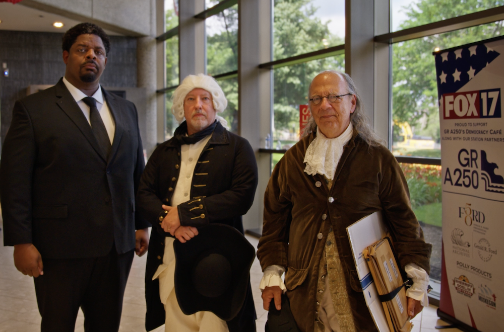 Three men, two in colonial costumes, one in a black suit, standing indoors by windows and a FOX 17 banner.
