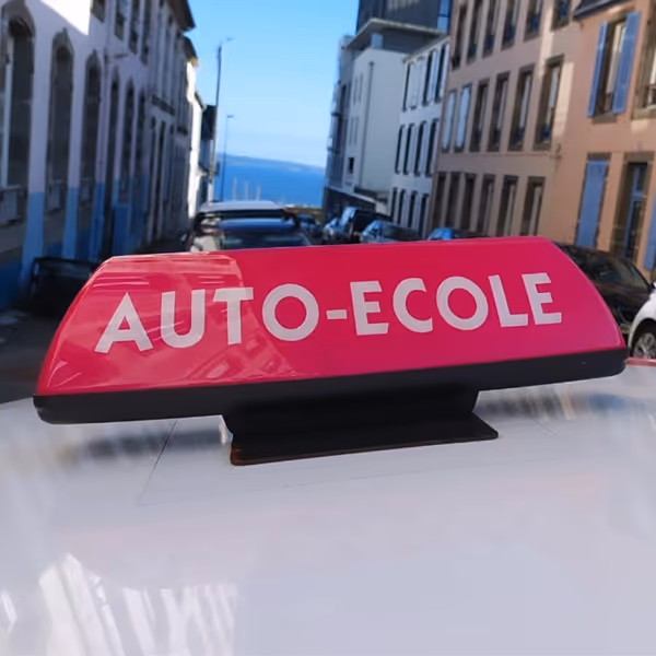 Signal lumineux rose marqué "AUTO-ÉCOLE" fixé sur le toit d’un véhicule d’apprentissage, en ville avec vue sur la mer en arrière-plan.