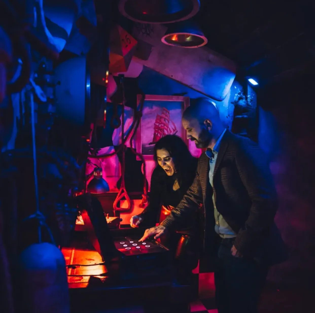 Two adults solving a puzzle on a touchscreen in a dimly lit, neon-themed escape room environment.
