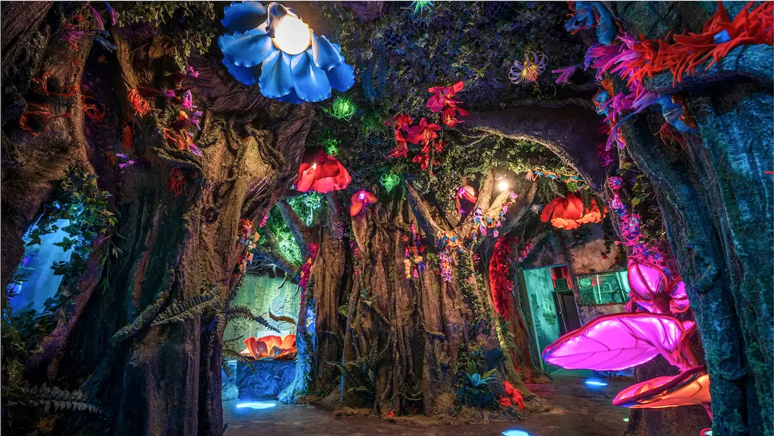 Whimsical indoor forest scene with glowing oversized flowers and twisted tree trunks, lit by colourful ambient lighting.