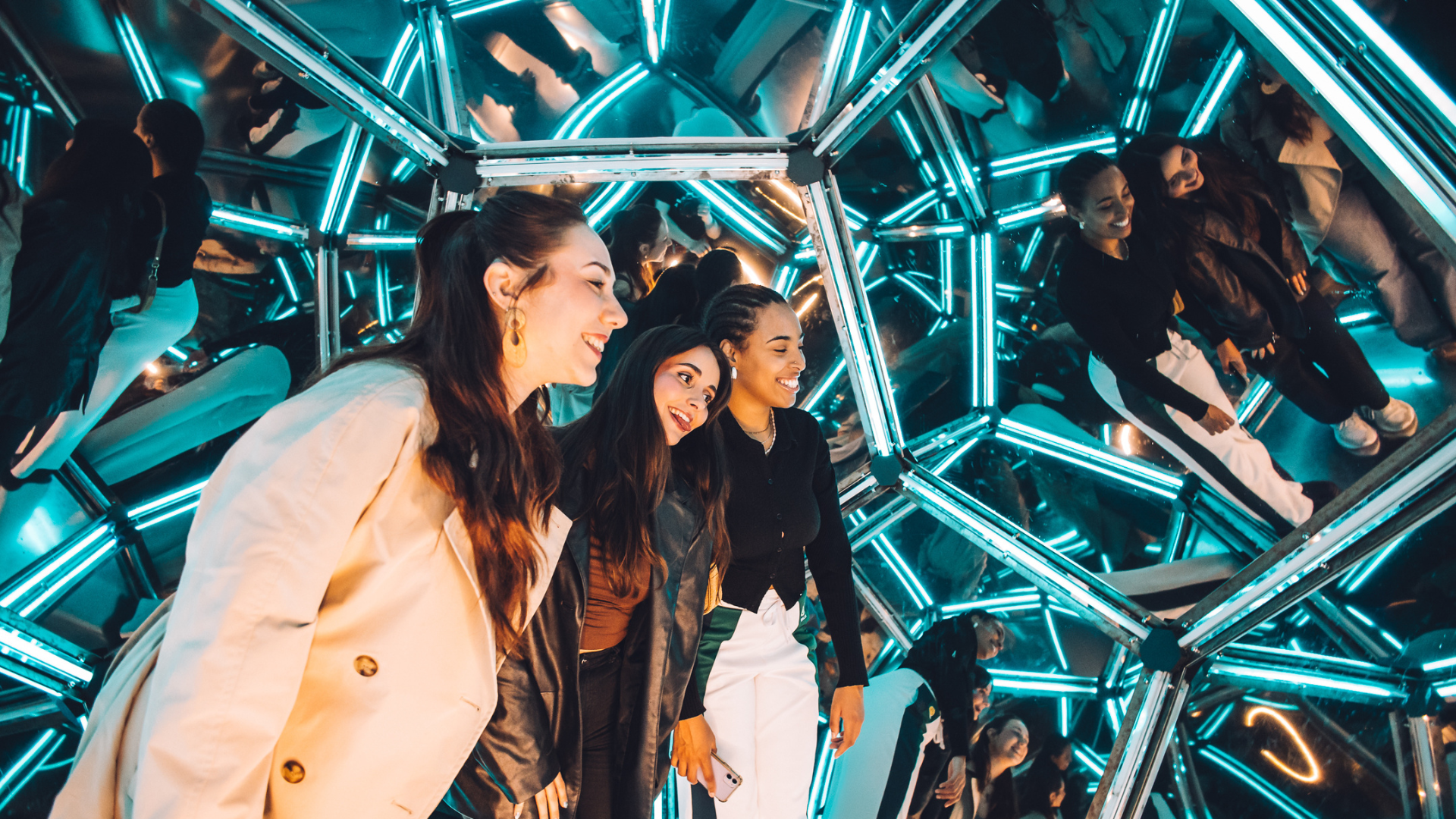 Three women smiling and posing inside a futuristic mirror room with glowing geometric light pattern