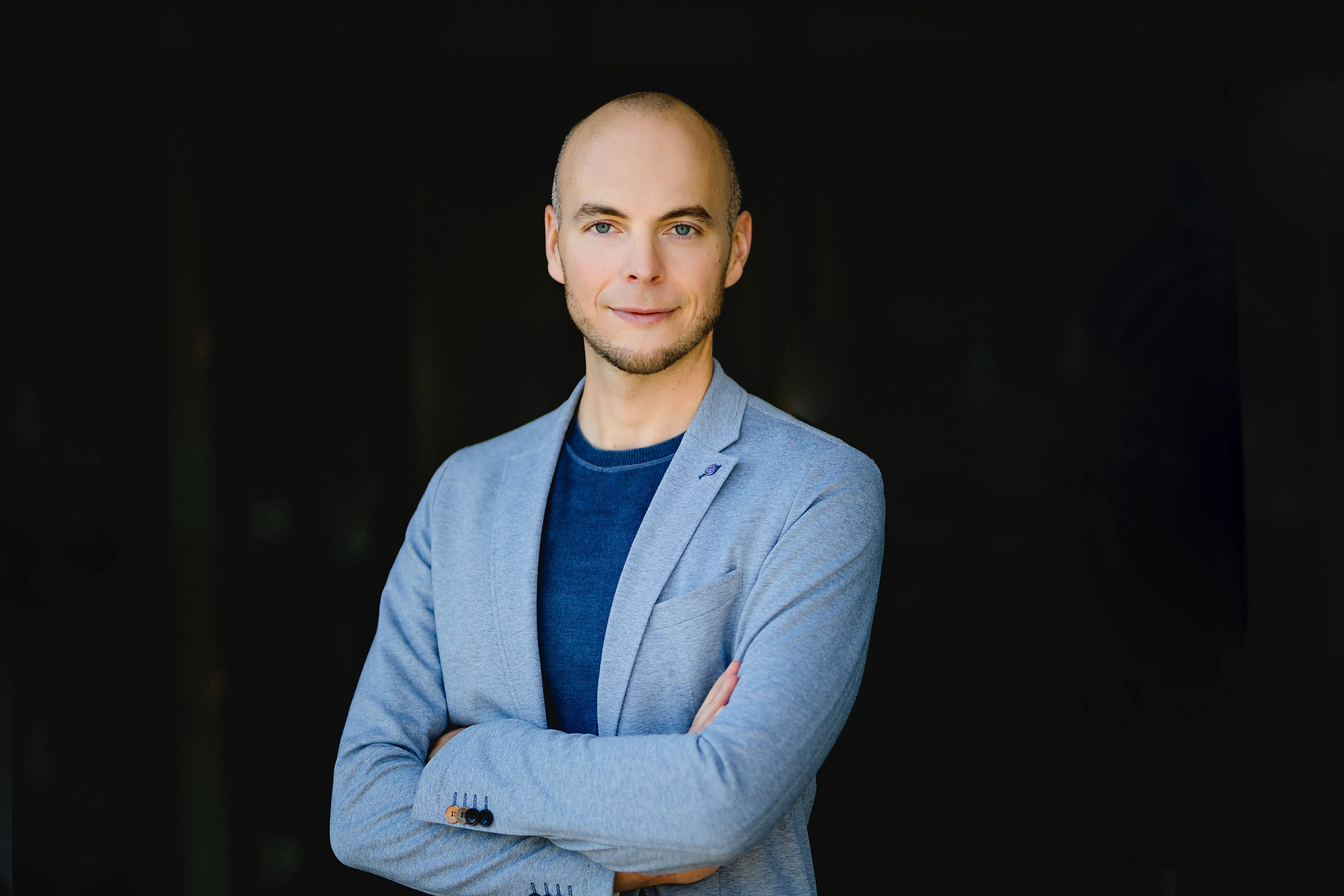Confident bald man with blue eyes wearing a light gray blazer and navy shirt standing with arms crossed against a dark background.