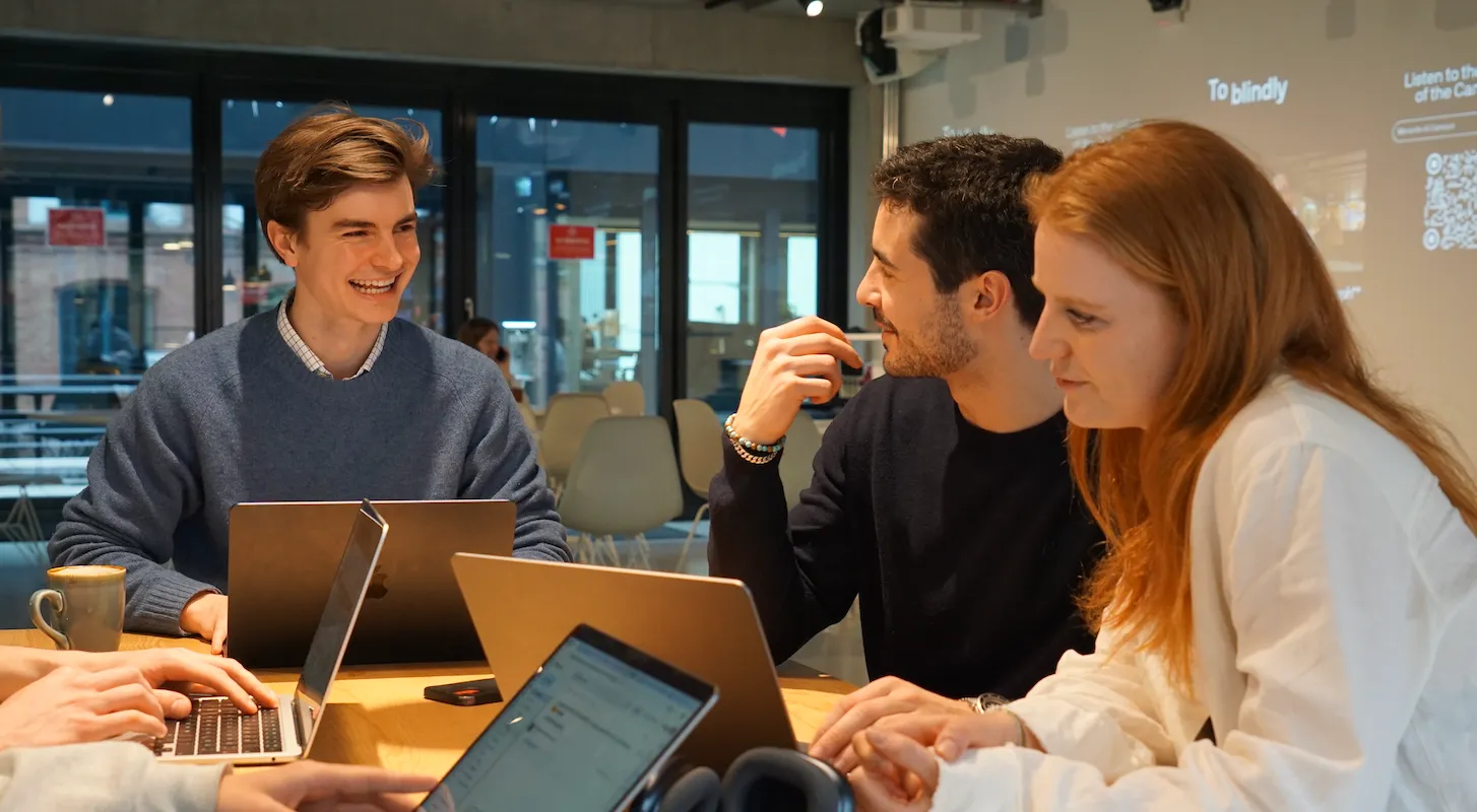 Three young adults sitting around a table with laptops, engaged in a friendly discussion in a modern office space.