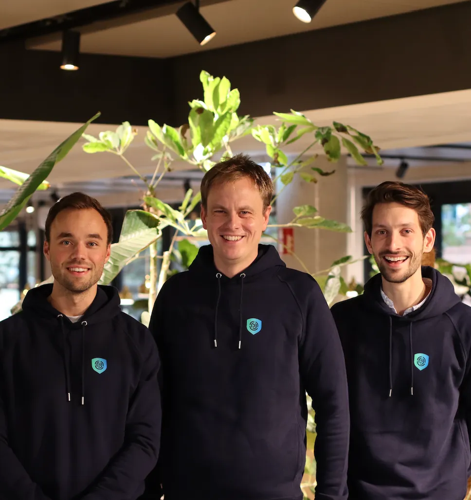 Three smiling men wearing matching black hoodies with a blue logo, standing indoors with green plants in the background.