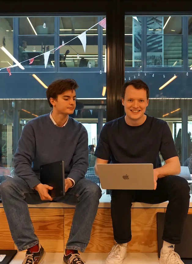 Two young men sitting on a wooden bench indoors, one holding a laptop and smiling, the other holding a folder and looking to the side.