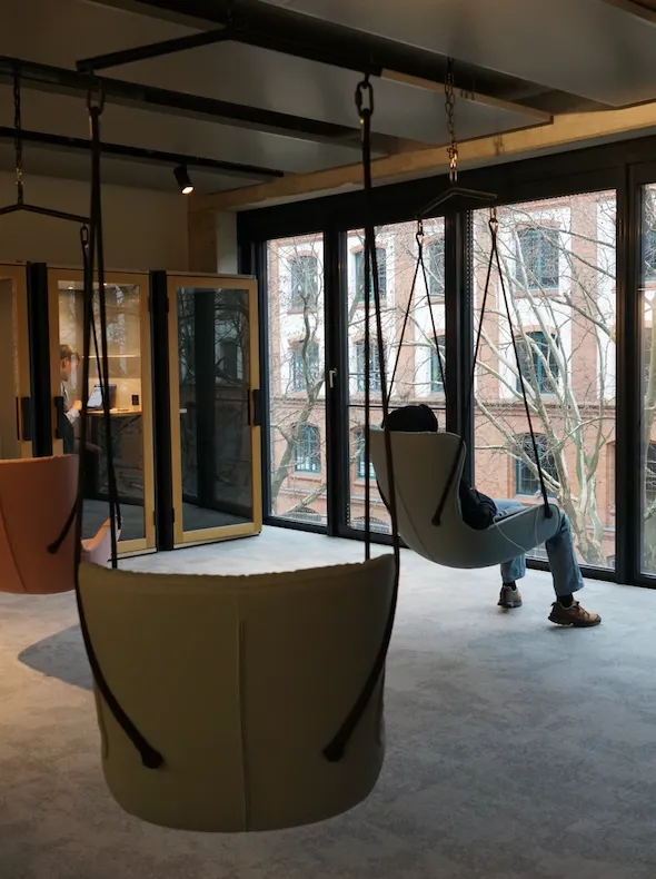 Modern office lounge with hanging chairs and large windows overlooking a brick building and leafless trees, with a person seated in one chair.