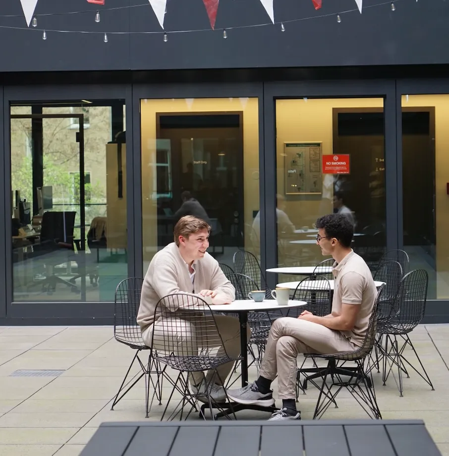 Two men sitting and conversing at an outdoor cafe table with coffee cups, in front of large glass doors.