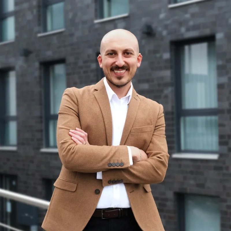 Smiling bald man in a brown blazer and white shirt standing with crossed arms in front of a modern building.