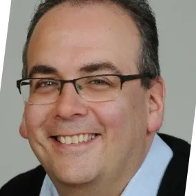 Smiling middle-aged man wearing glasses and a light blue shirt with a dark jacket.