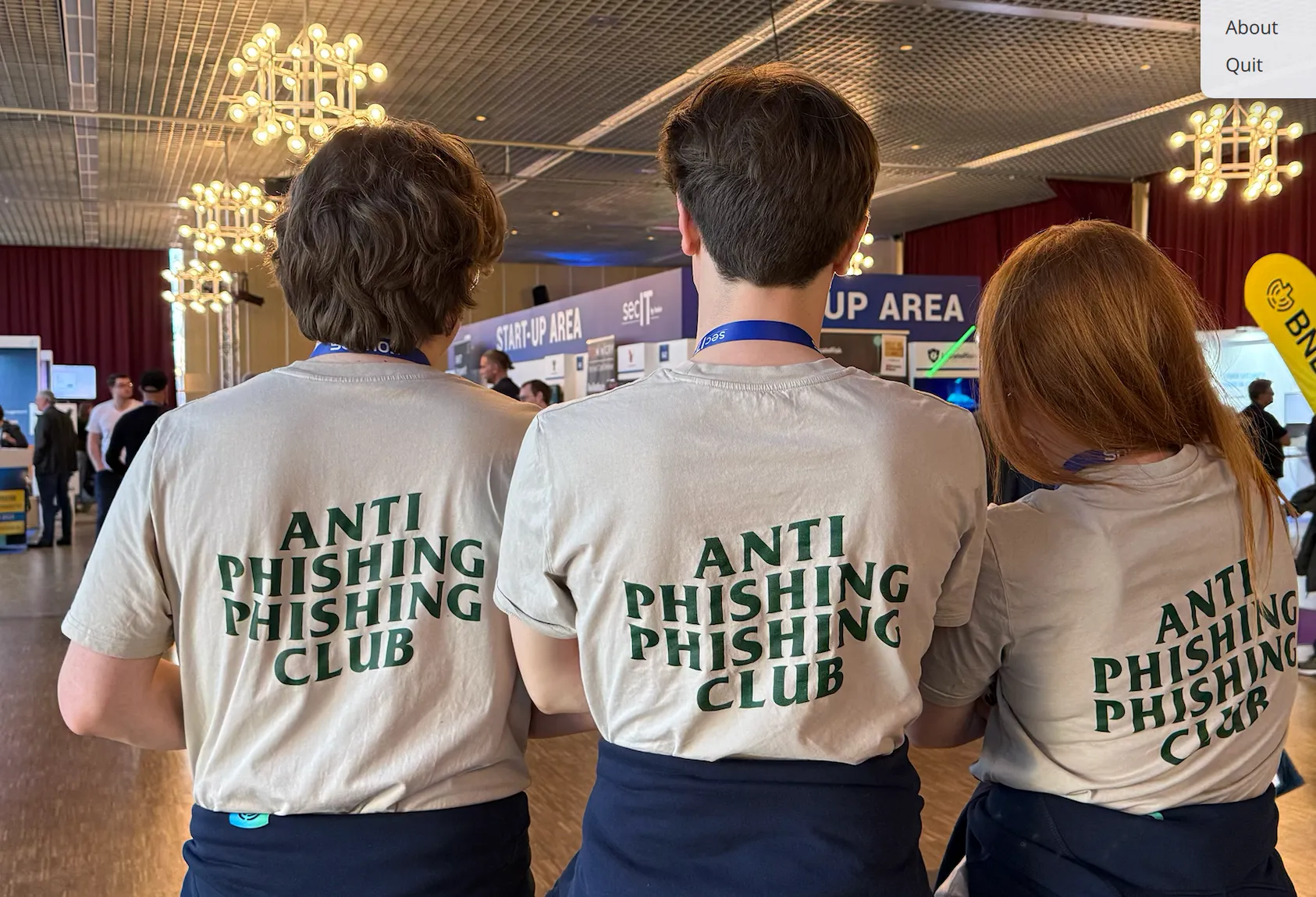 Three people standing indoors with their backs to the camera wearing matching beige t-shirts that say 'ANTI PHISHING PHISHING CLUB.'