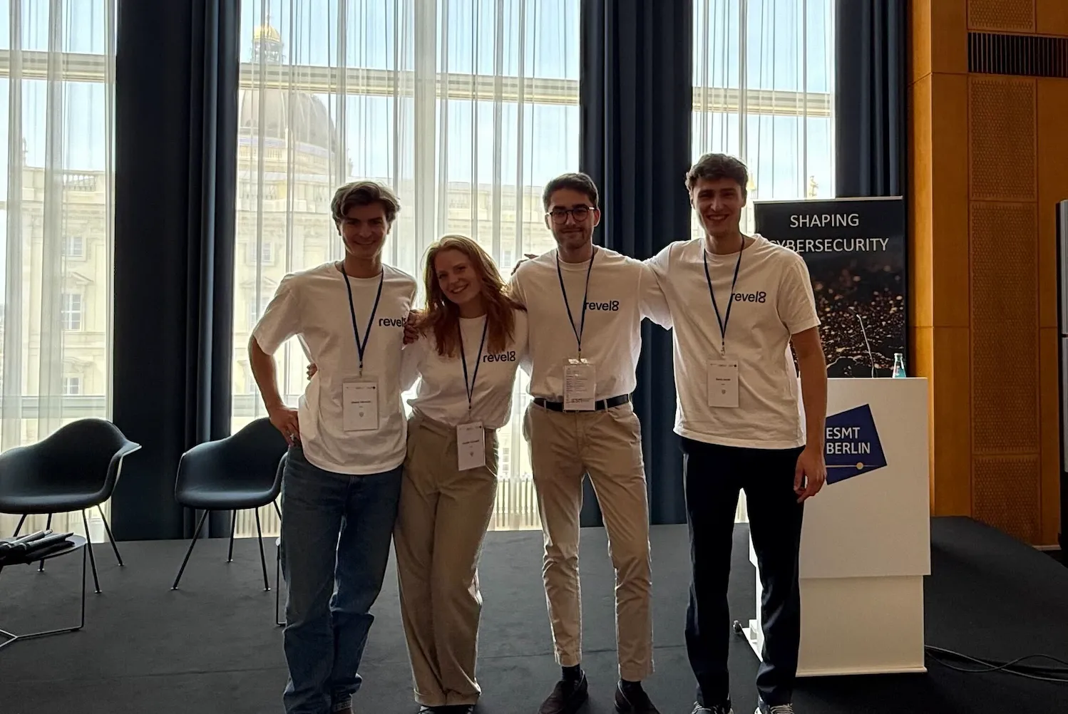 Four young adults in matching white revel8 t-shirts standing arm-in-arm in front of large windows and a podium with ESMT Berlin branding.