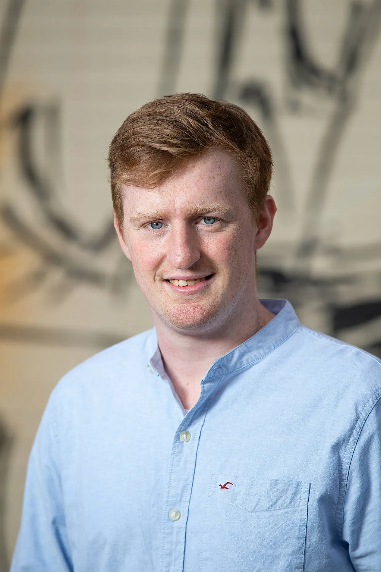 Smiling young man with red hair, freckles, and blue eyes wearing a light blue button-up shirt.