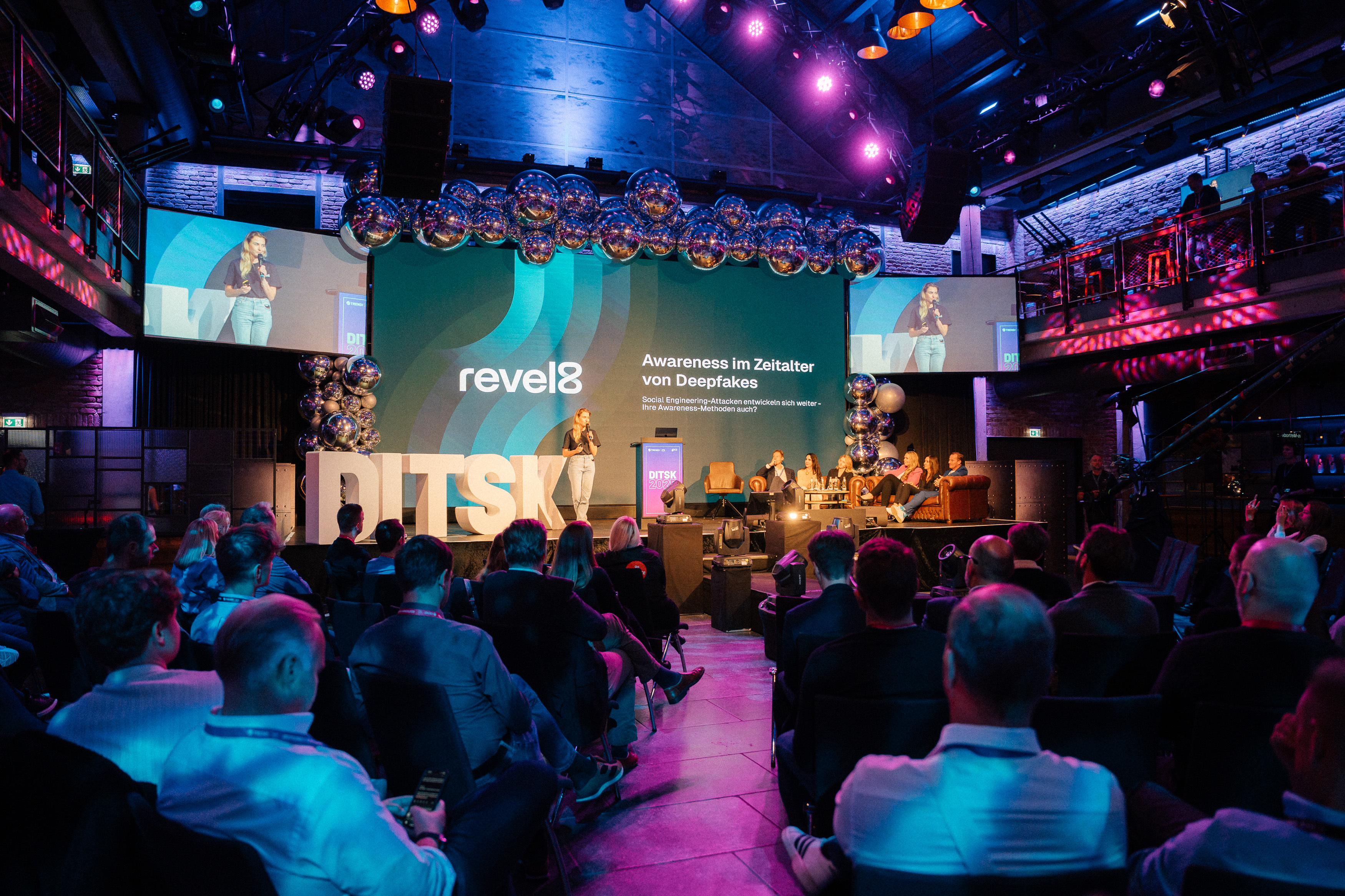 Woman speaking on stage at a conference with audience seated, large screen behind displaying 'revel8' and text on deepfake awareness.