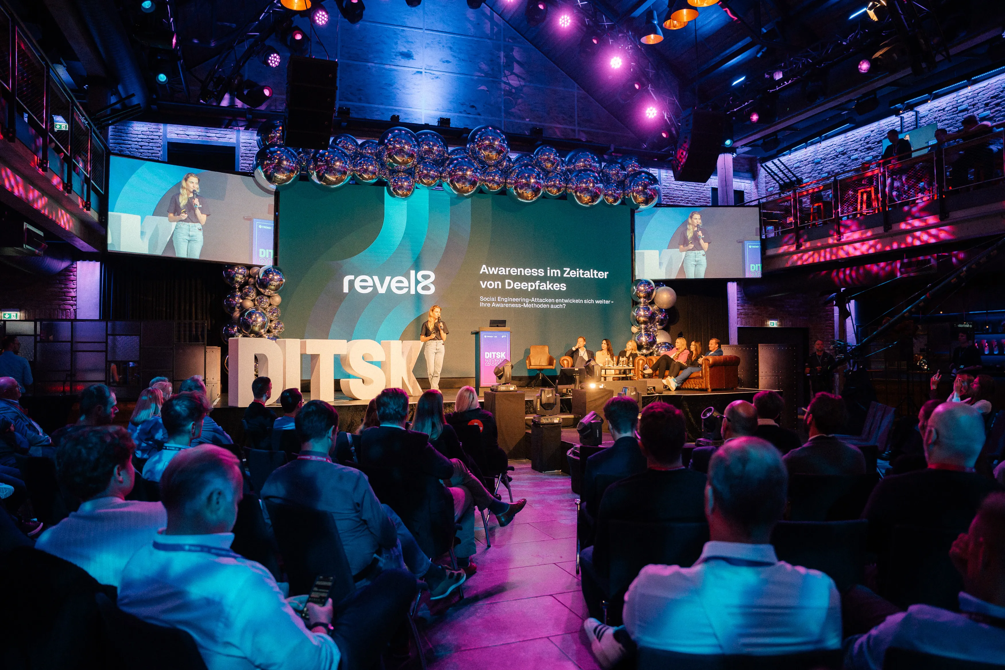 Woman speaking on stage at a conference with audience seated, large screen behind displaying 'revel8' and text on deepfake awareness.