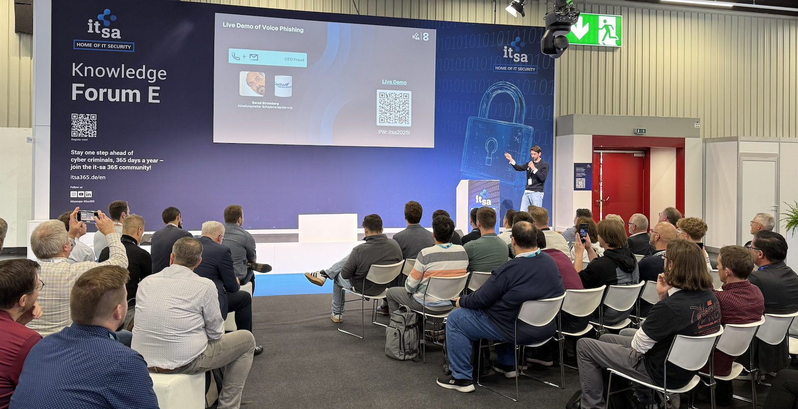 Audience seated in a conference room watching a presenter demonstrate voice phishing on a large screen at the it-sa Knowledge Forum E.