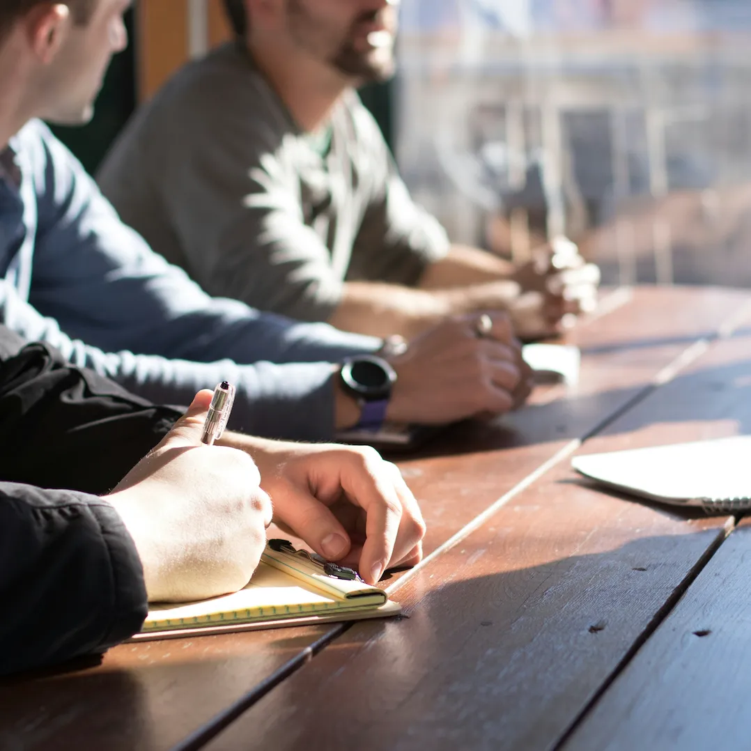 People sitting at a wooden table, one person writing notes on a yellow notepad with a pen.