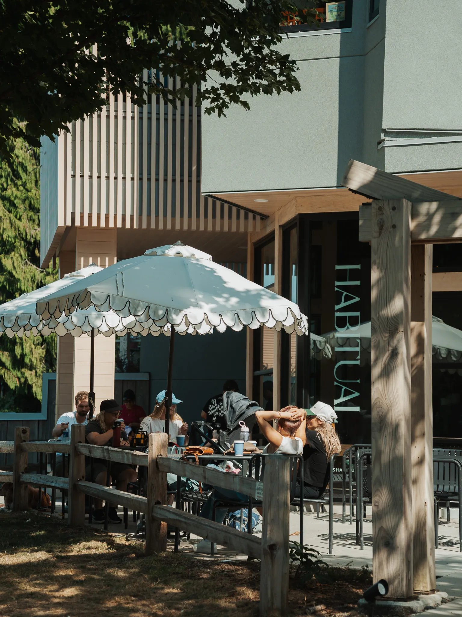 Outdoor cafe seating area with people sitting at tables under white scalloped umbrellas, next to a modern building with a wooden fence and trees in the background.