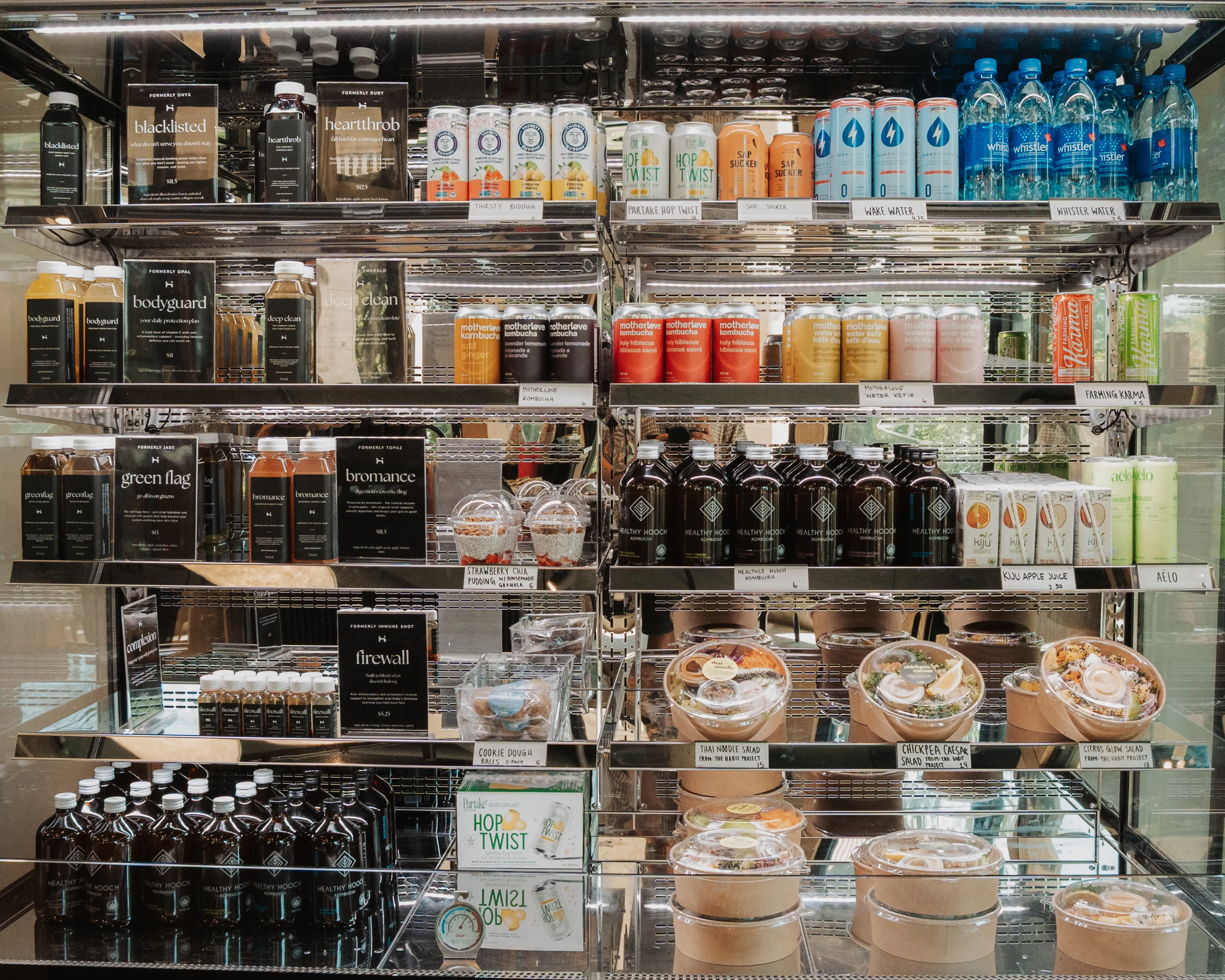 Refrigerated display case with shelves stocked with a variety of beverages including bottled water, kombucha, juices, and canned drinks, along with packaged salads and desserts labeled with prices.