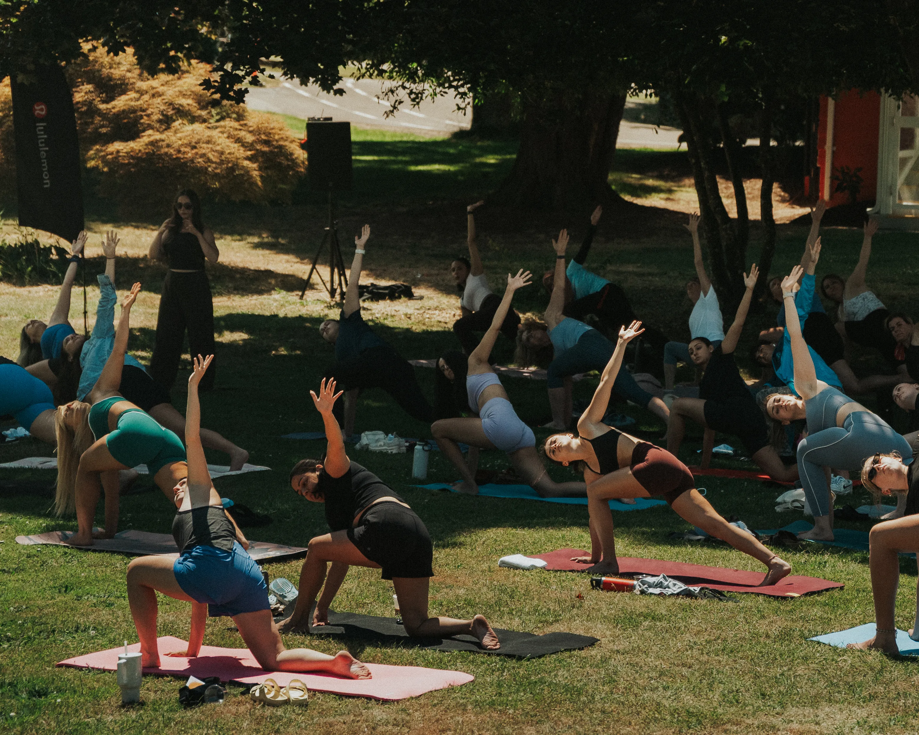 Group of people practicing yoga outdoors on mats in a grassy park area, all performing a side stretch pose with one hand on the ground and the other reaching up, while an instructor stands nearby observing.
