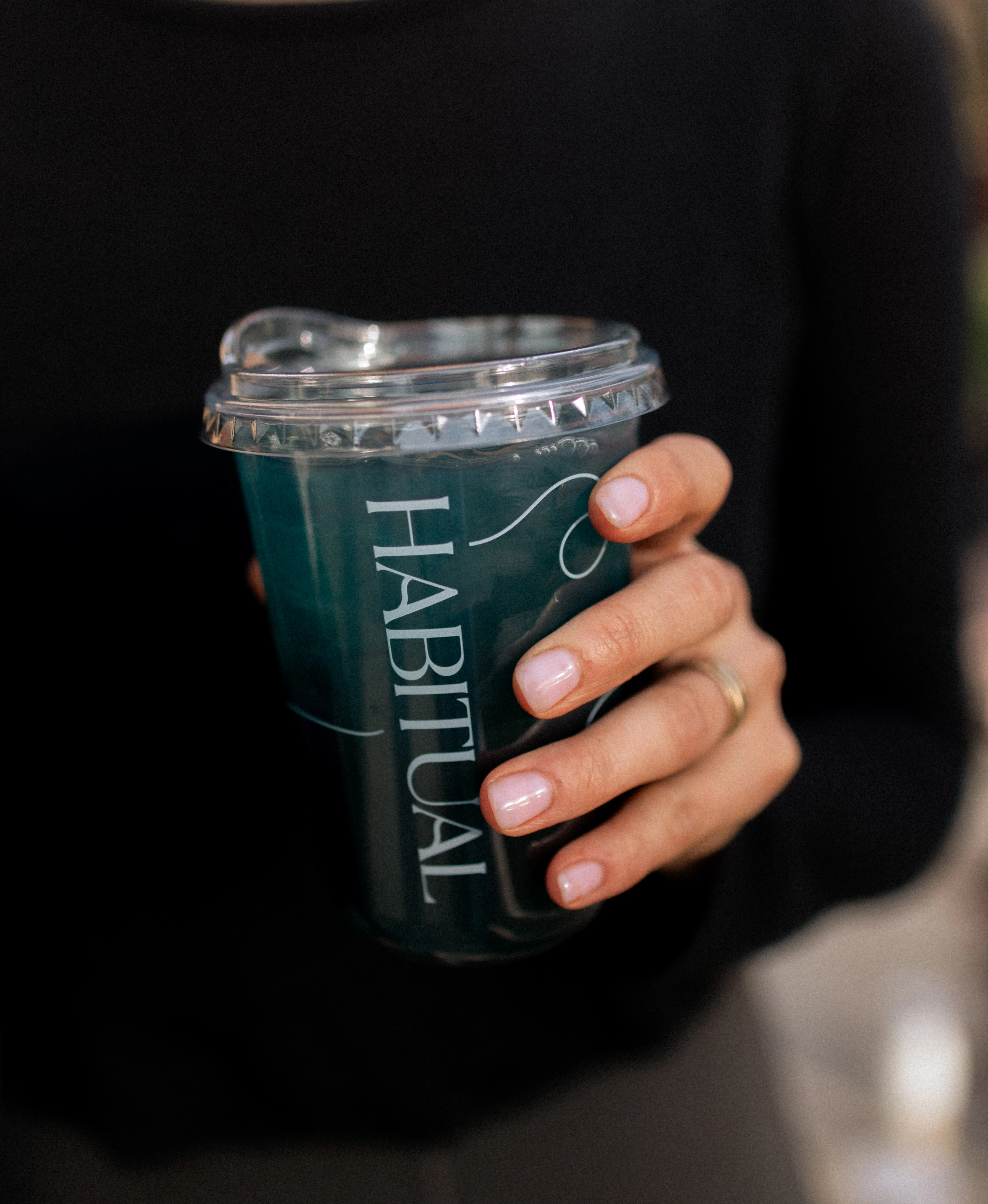 Hand with polished nails and a gold ring holding a plastic cup of dark beverage with a clear lid, the cup labeled HABITUAL in white script.