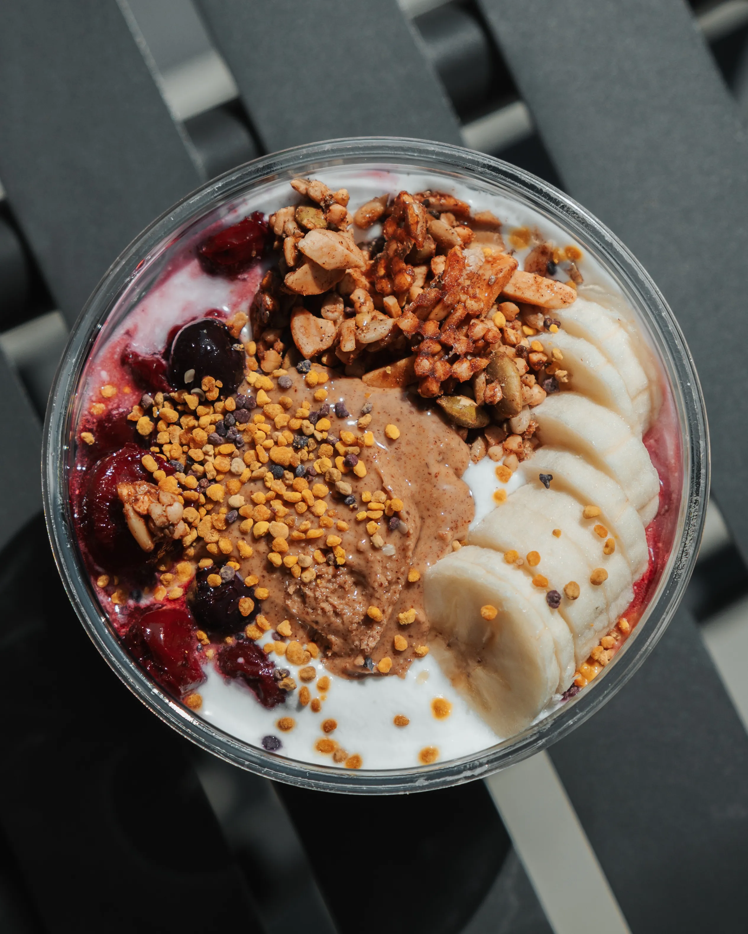 Overhead view of a clear bowl containing yogurt topped with sliced bananas, almond butter, granola, dark berries, and sprinkled bee pollen.