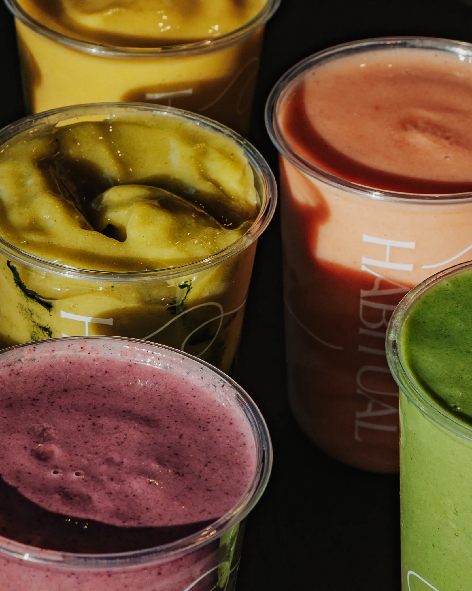 Close-up of five plastic cups filled with colorful smoothies in yellow, green, purple, pink, and orange hues on a dark surface.