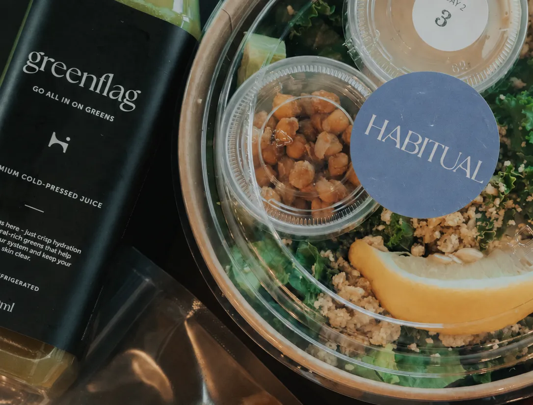 Black bottle of Greenflag cold-pressed juice next to a salad bowl with chickpeas, kale, lemon slice, and a blue Habitual sticker on the lid.
