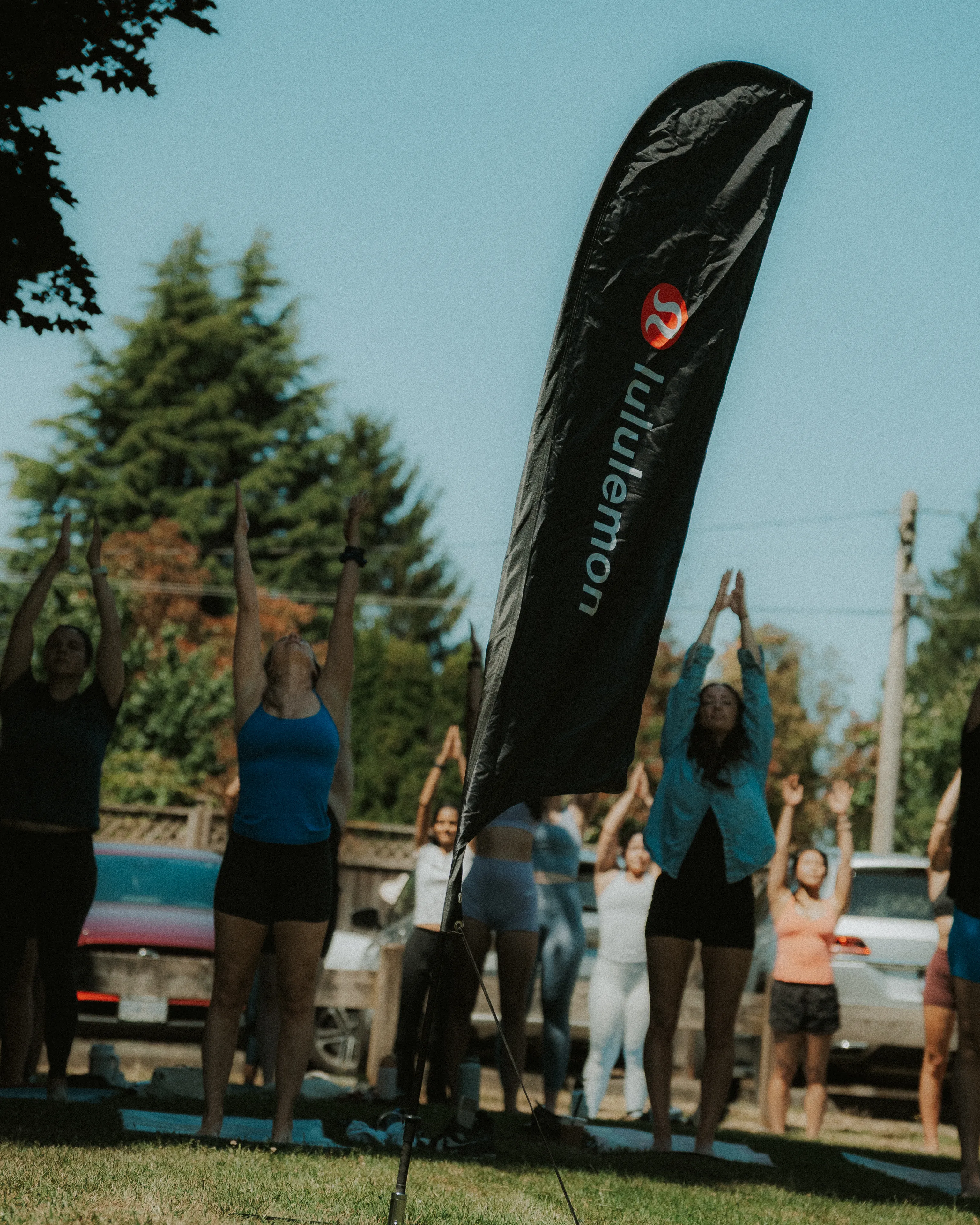 Group of people outdoors practicing yoga with arms raised, next to a black vertical flag with the lululemon logo on a grassy area with trees and parked cars in the background.