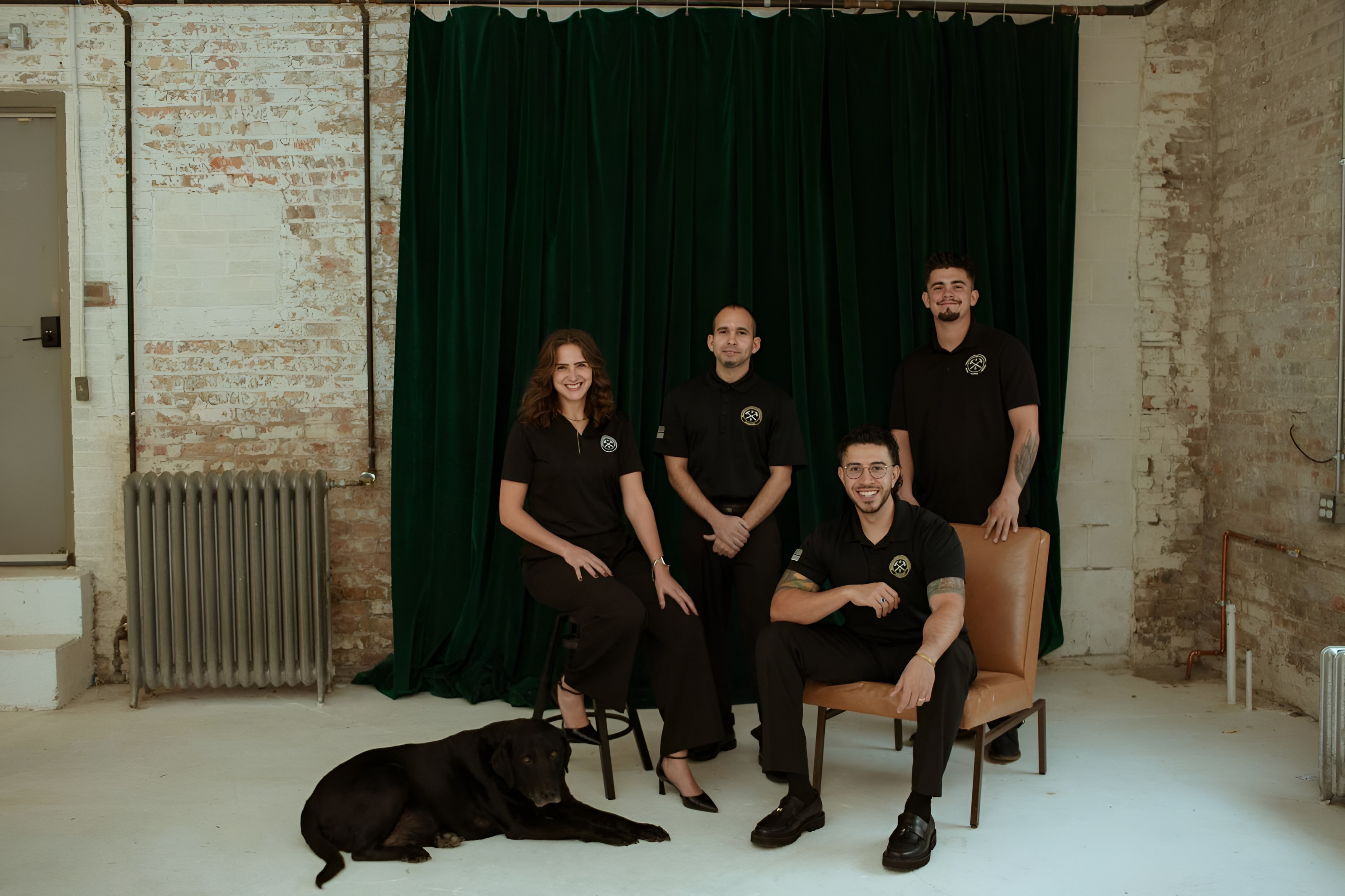 Four people in black uniforms posing indoors with a black dog lying on the floor in front of them against a green curtain and exposed brick wall.