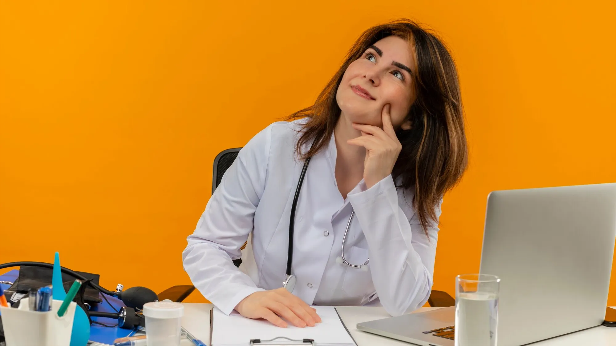 A female doctor in a white coat with a stethoscope sits at a desk with a laptop and medical tools, smiling thoughtfully with her finger on her cheek, against a bright orange background.