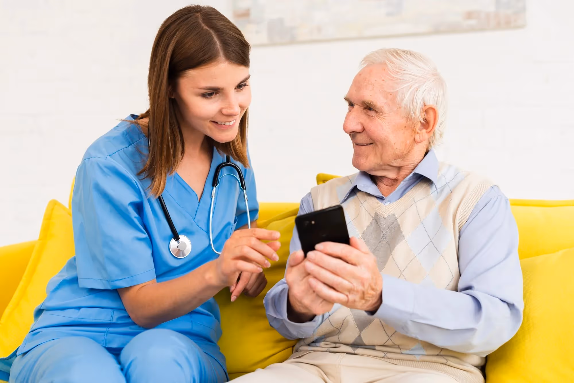 A young female nurse in blue scrubs with a stethoscope is sitting next to an elderly man on a yellow couch. She is smiling and pointing at a smartphone he is holding, as they both look at the screen together.