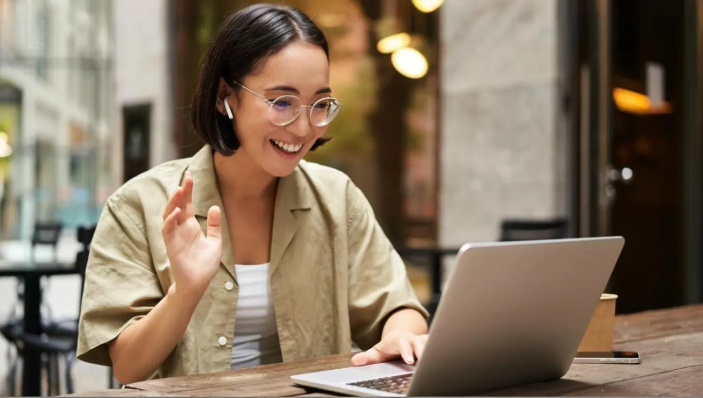 A woman wearing wireless earbuds smiles and waves at her laptop during a video call in an outdoor café setting. The scene reflects remote work or virtual meetings using Teams for business. Keywords: Microsoft Teams calling, Teams for business, Teams contact center integration.