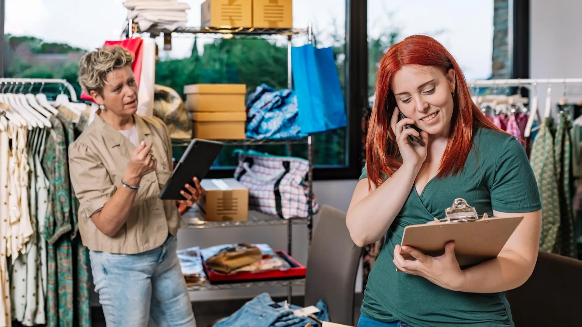Two women in a retail clothing store collaborate—one speaks on the phone holding a clipboard, while the other gestures with a tablet. Surrounded by clothing racks and packages, they represent a small business using cloud-based retail communication solutions to manage inventory and coordinate customer service.
