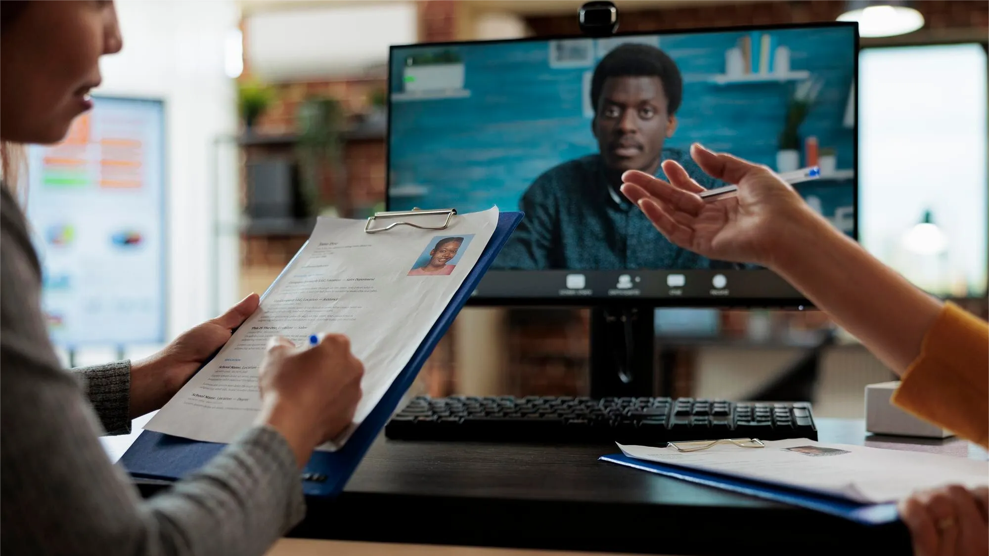 Two people sit at a desk with printed resumes and documents, conducting a remote video interview with a man shown on a monitor. The image illustrates virtual hiring or collaboration powered by Teams contact center integration and Microsoft Teams calling. Keywords: Teams for business, Microsoft Teams calling, Teams contact center integration.