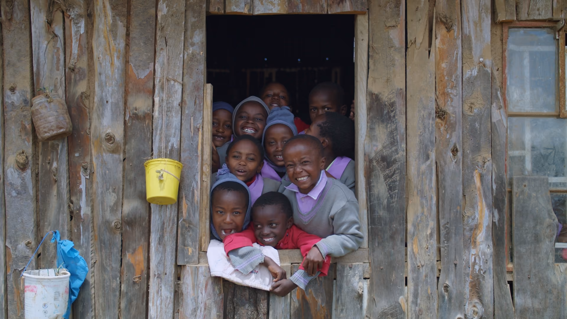 Group of smiling children peering out a rustic wooden window of a building.