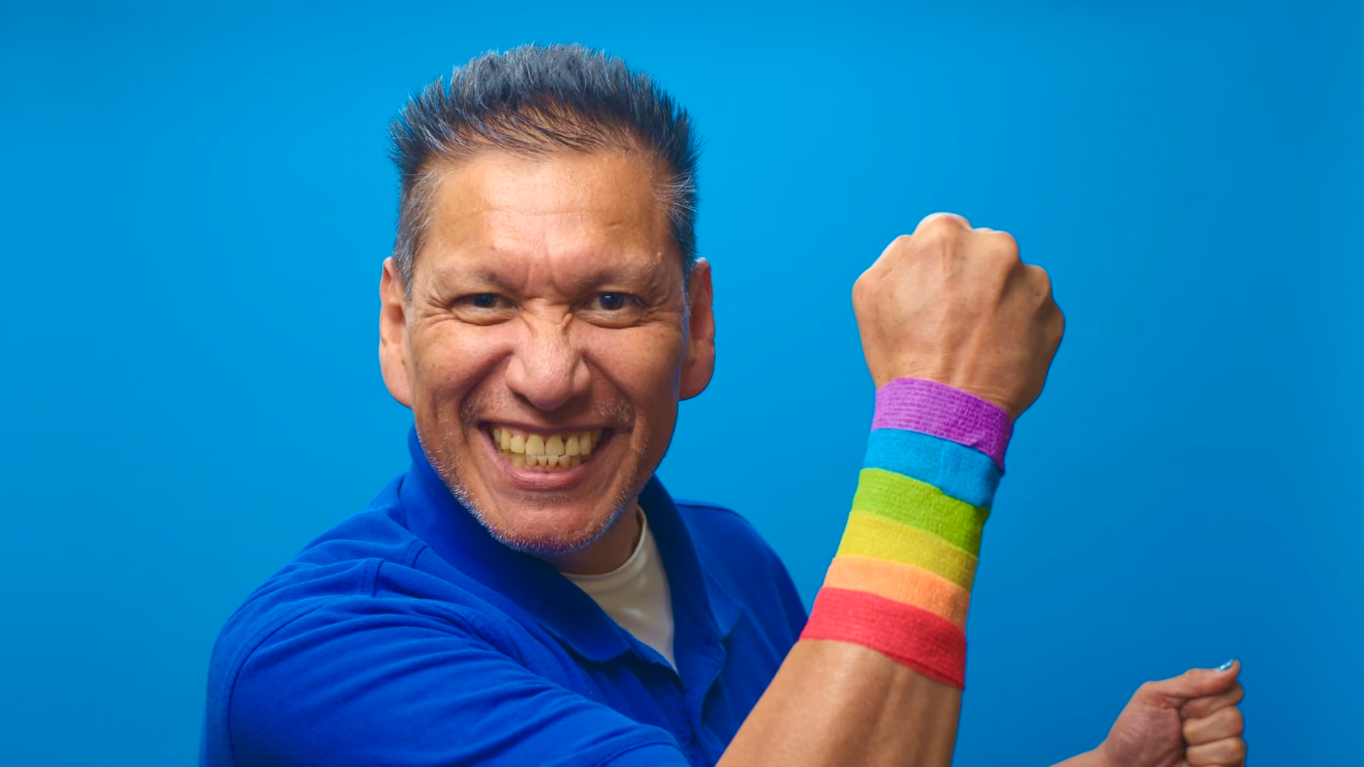 Smiling man raising fist with a rainbow wristband on blue background celebrating pride.