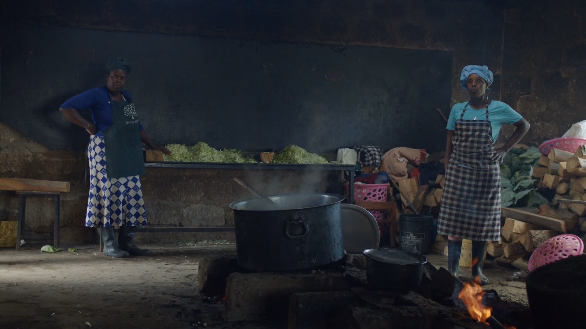 Two women in aprons standing near a large steaming pot over an open fire in a rustic kitchen.
