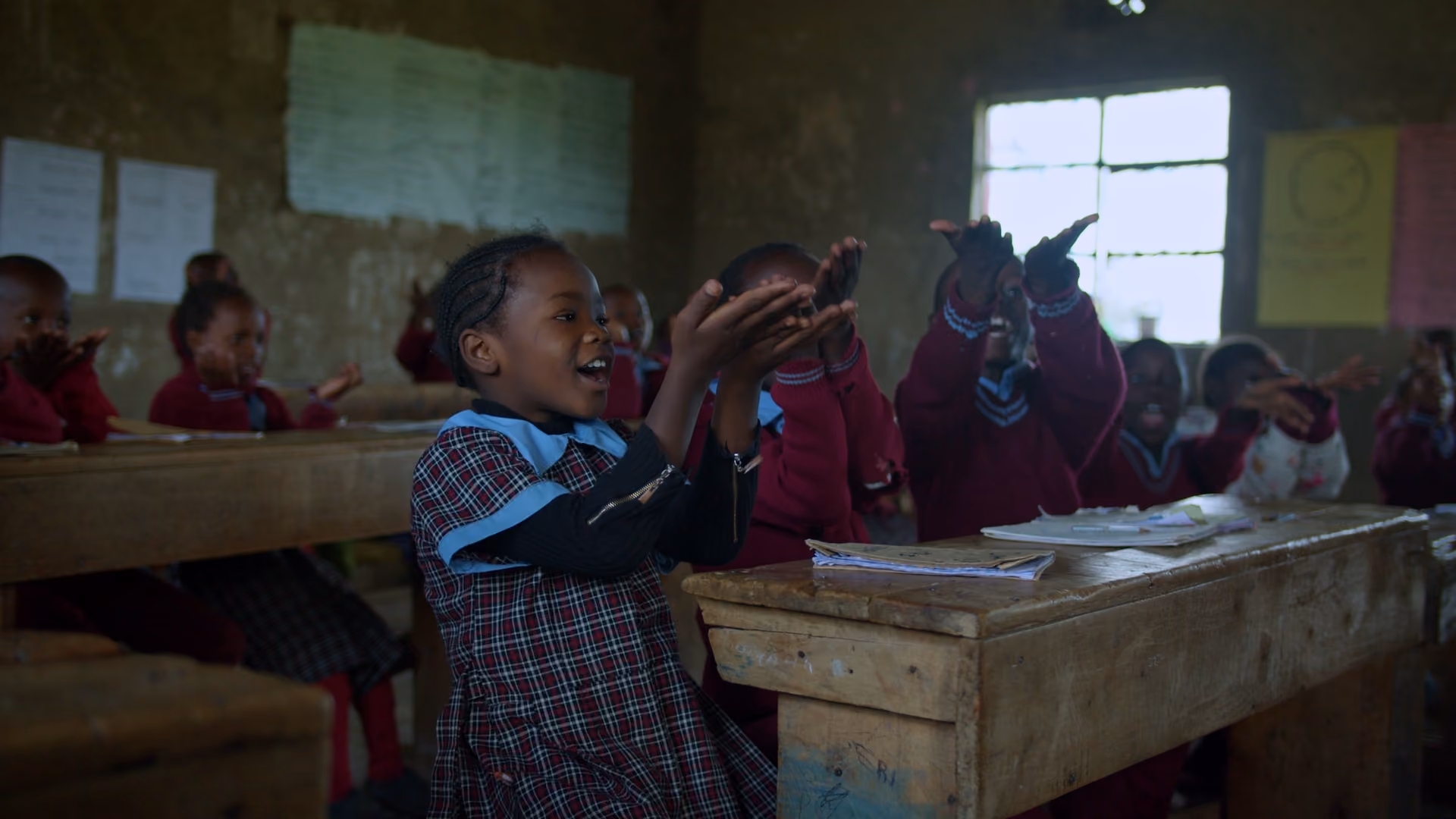 Children in a crustic lassroom clapping and smiling while sitting at wooden desks with notebooks.