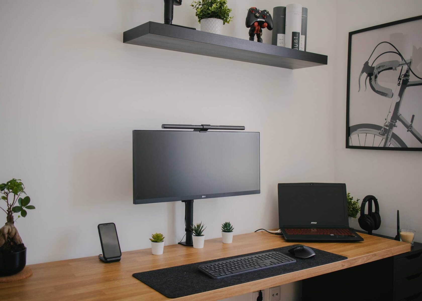 A desk featuring a laptop, keyboard, monitor, and a shelf above with various plants.