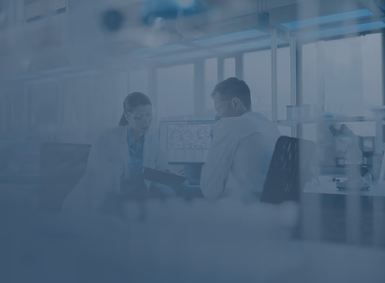 Two scientists in lab coats having a discussion in a laboratory with scientific equipment and monitors in the background.