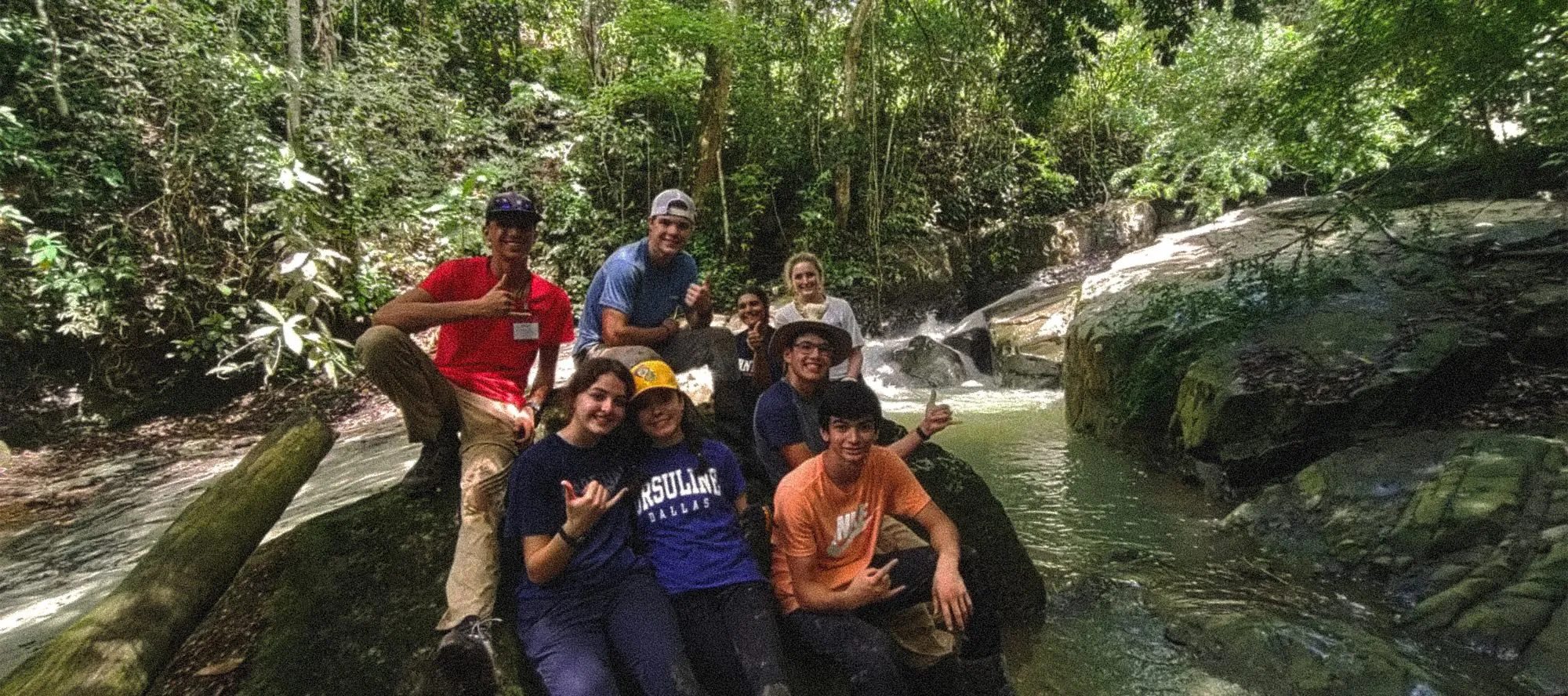 Group of young people enjoying a nature adventure in a tropical rainforest, sitting by a waterfall surrounded by lush greenery.